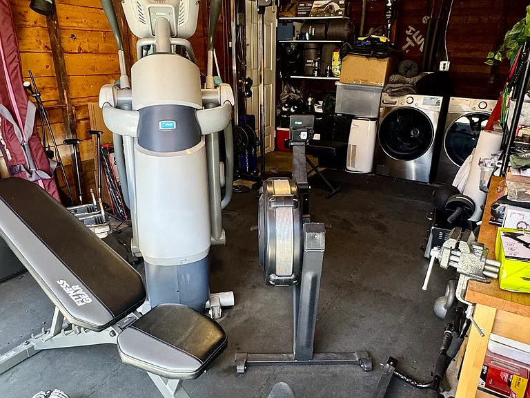 Rubber mat covers floor. All equipment is Commercial grade incl. AMT elliptical, Olympic rower, adjustable weight bench & dumbbells, wall-anchored pulley system with Lat bar, rope & hand tow triangles. Wall mounted 43" inch flat screen TV on the right.
