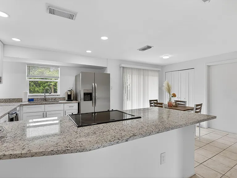 Open kitchen with a large granite island, stainless refrigerator, and adjacent dining area with sliding glass doors.
