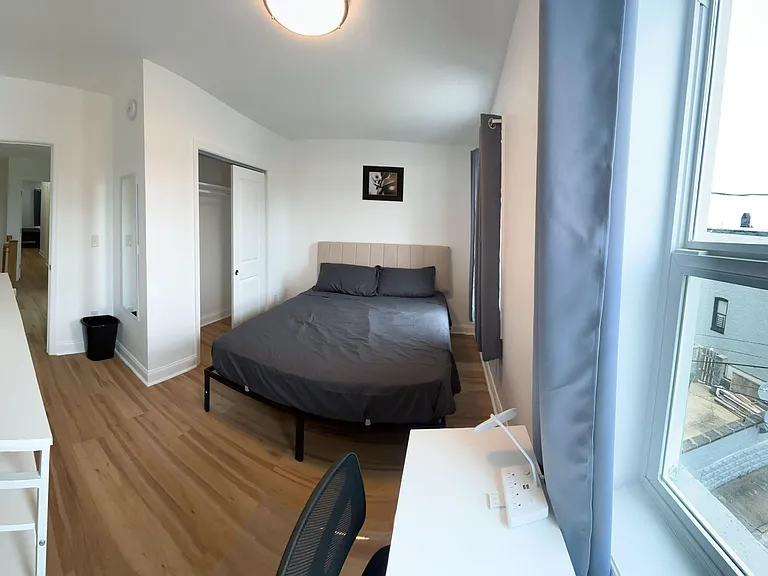Furnished bedroom with a full bed, large window with curtains, and a desk setup, plus an open closet for storage.