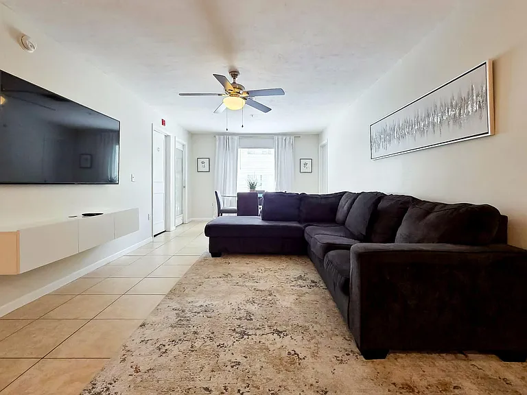 Open living area with sectional seating, wall-mounted TV, and ceiling fan, flowing into a dining nook by the window.