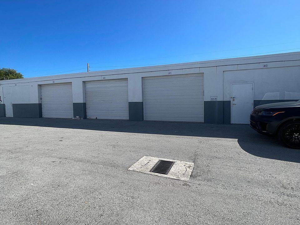 Front exterior view of warehouse unit with roll-up metal door, side access door, paved driveway, and clear sunny surroundings.