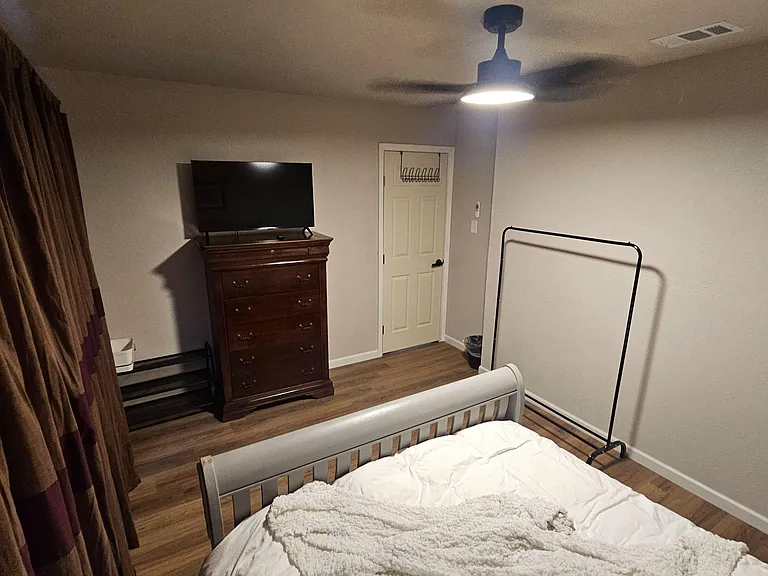 Bedroom with wood-look flooring, ceiling fan light, dresser with TV, and space for a clothing rack or extra storage.