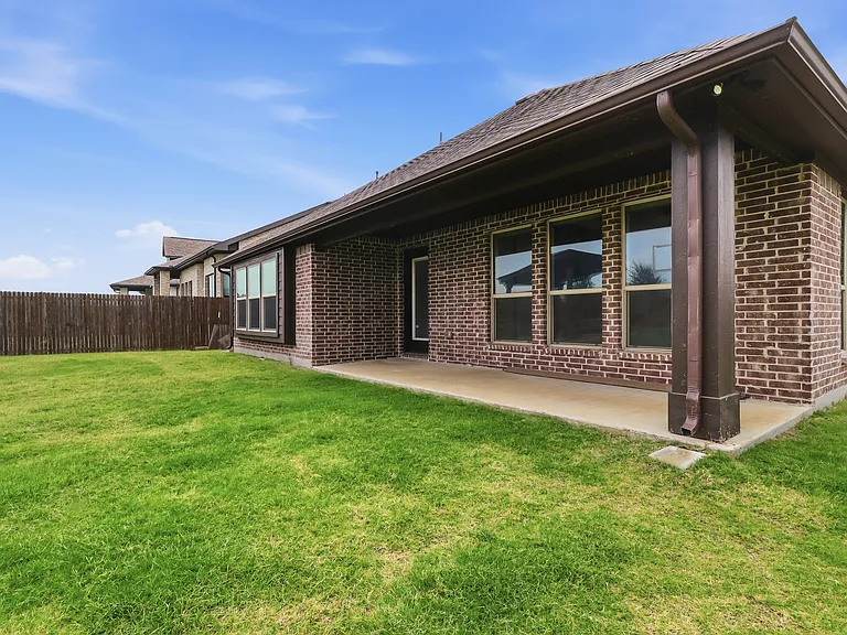 Expansive green lawn featuring a covered patio, brick exterior, multiple windows, and a wood privacy fence