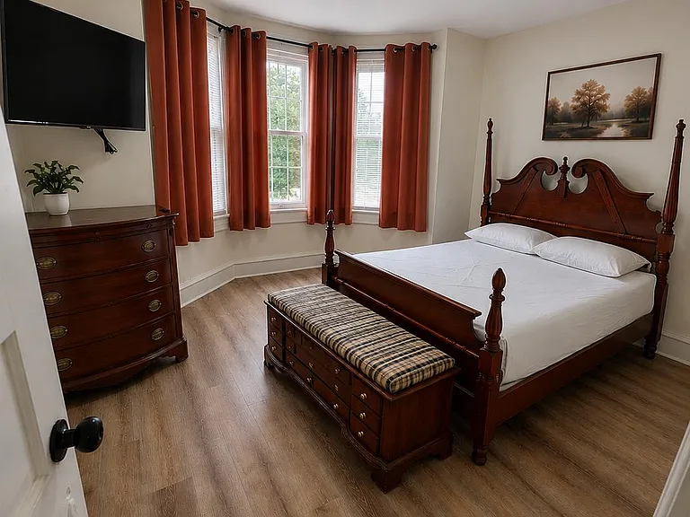 Furnished bedroom featuring a classic wood bed, dresser, and bench, plus a wall-mounted TV and bay windows with curtains.