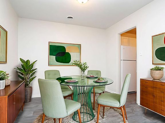 Chic dining area with a modern glass table, stylish green chairs, vibrant art, and sleek wooden cabinet, offering a cozy and inviting atmosphere.