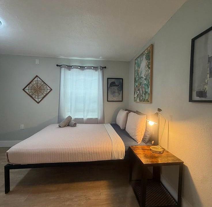 Furnished bedroom with a full/queen bed, nightstand with lamp, and a window with curtains for natural light.