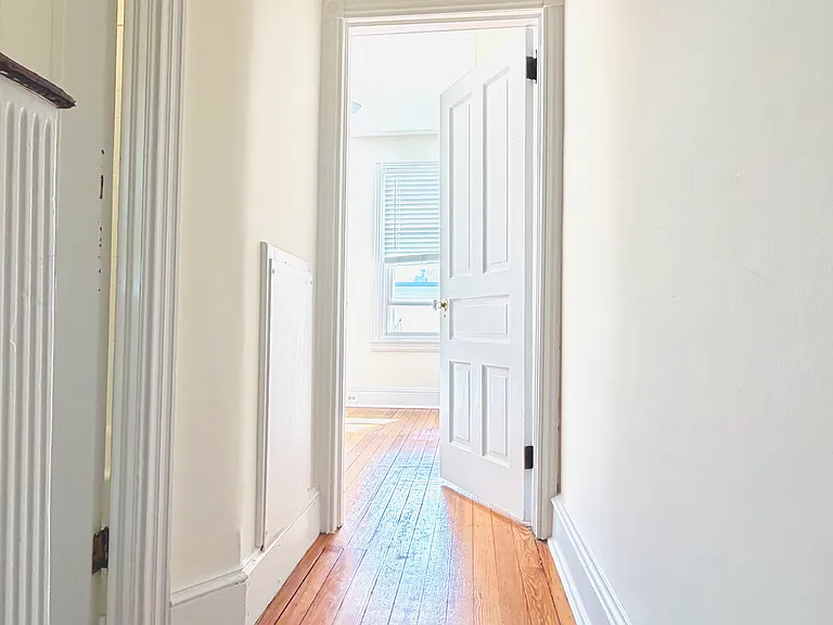 Second-floor hallway with original hardwood floors, wall sconce, decorative trim, and radiator leading to a sun-lit bedroom.