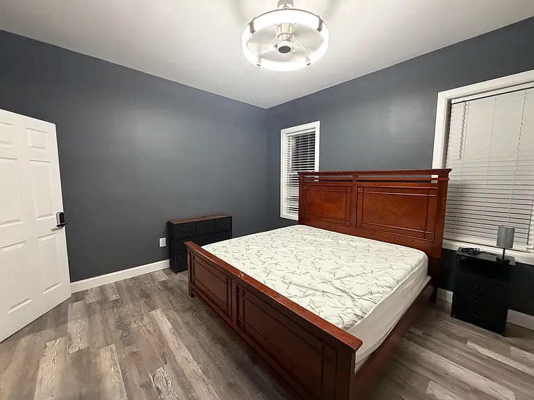 Private bedroom with wood-style flooring, a large bed frame, and two windows with blinds for light and privacy.