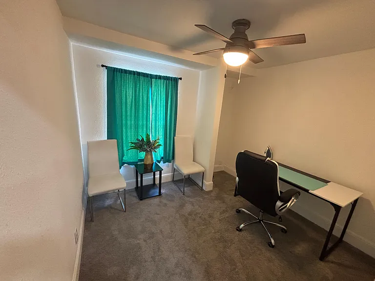 Carpeted office space with ceiling fan, window with curtains, and room for a desk or small seating area.