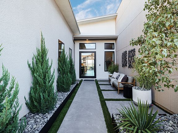 A narrow, paved walkway flanked by lush greenery, including tall cypress trees and other potted plan