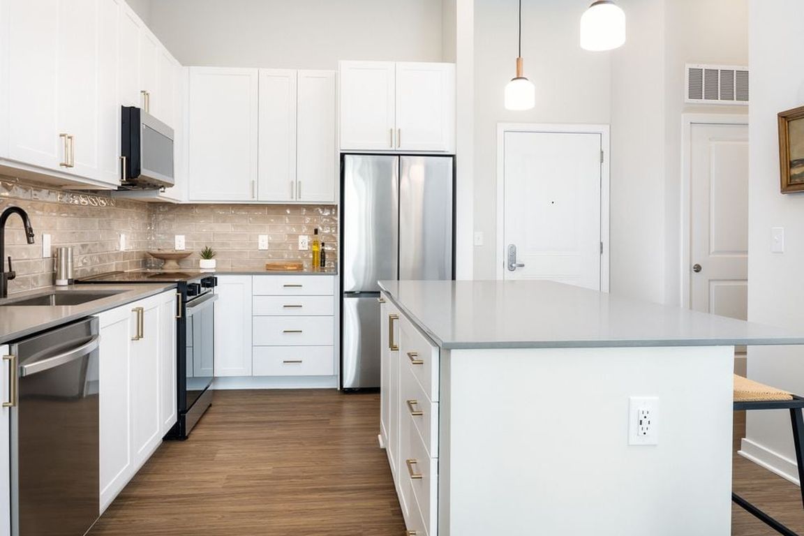 Upgrade Collection kitchen with white cabinetry, beige tile backsplash, grey quartz countertops, pendant lighting, and hard surface vinyl plank flooring (Representative photo)