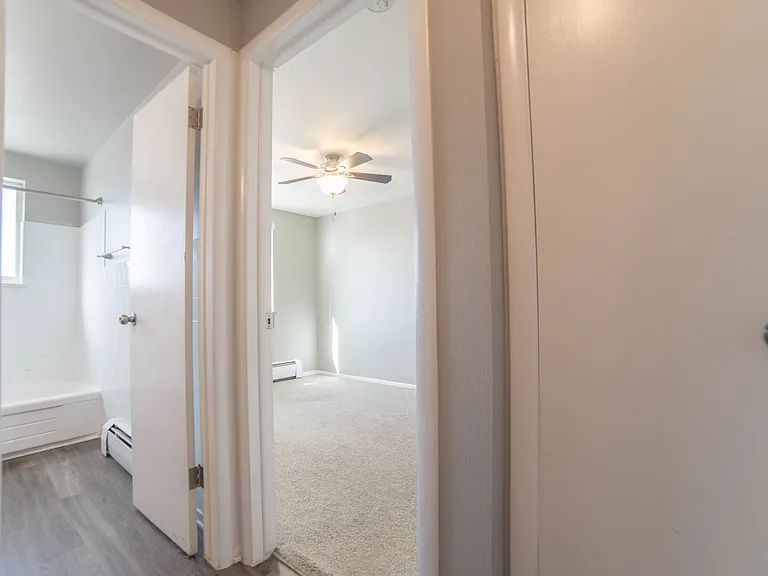 Inviting hallway view showcasing a bright and airy bedroom with soft carpet and a ceiling fan, adjacent to a modern bathroom with sleek floors.