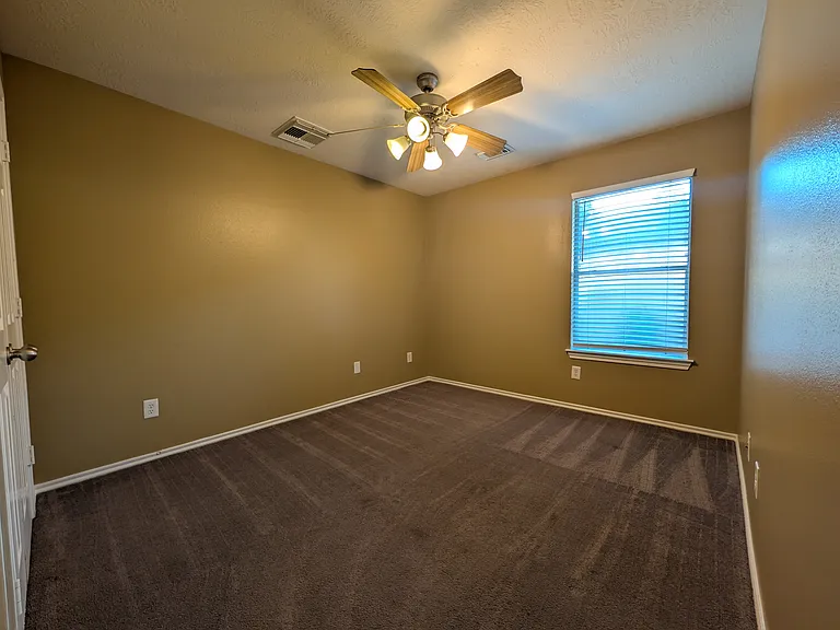 This bright guest bedroom offers plush carpeting, neutral tan paint, and a large window with blinds for ample natural light.