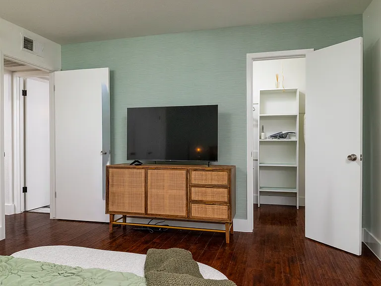 Rest in a calming bedroom with soft green tones, warm lighting, and a cozy bench at the foot of the bed. Enjoy a large TV, stylish dresser, and easy access to a spacious walk-in closet for a relaxing, comfortable stay.
