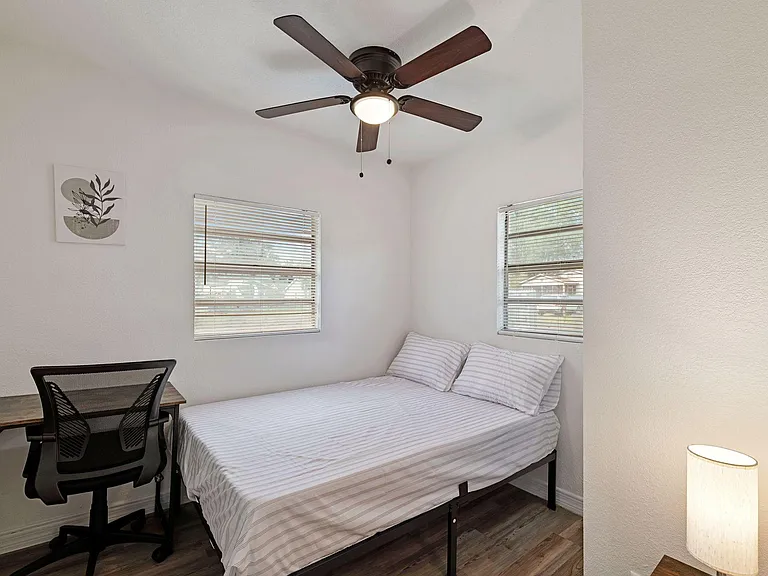 Furnished bedroom with a full-size bed, desk workspace, ceiling fan, and dual windows with blinds for natural light.