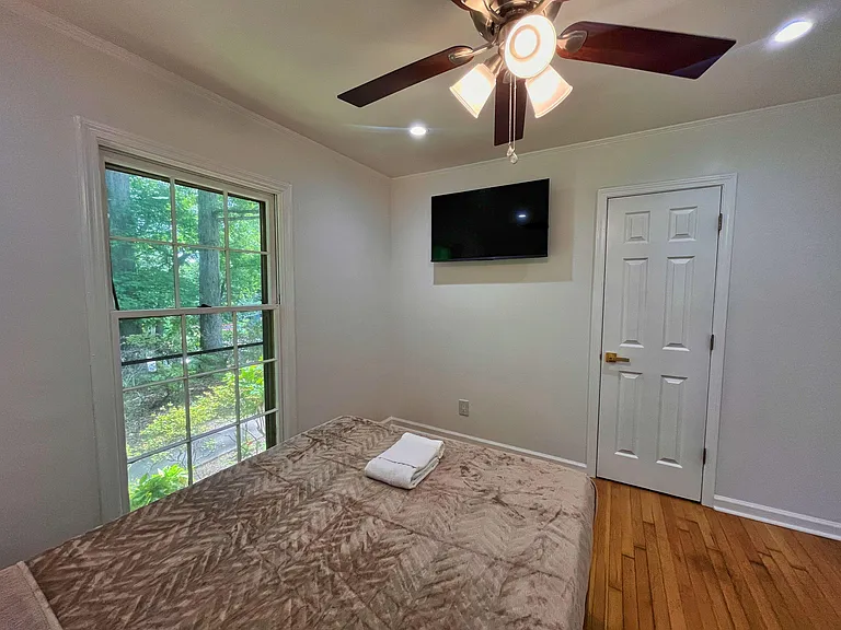 Private bedroom with hardwood floors, a large window with leafy views, ceiling fan, and a wall-mounted TV.