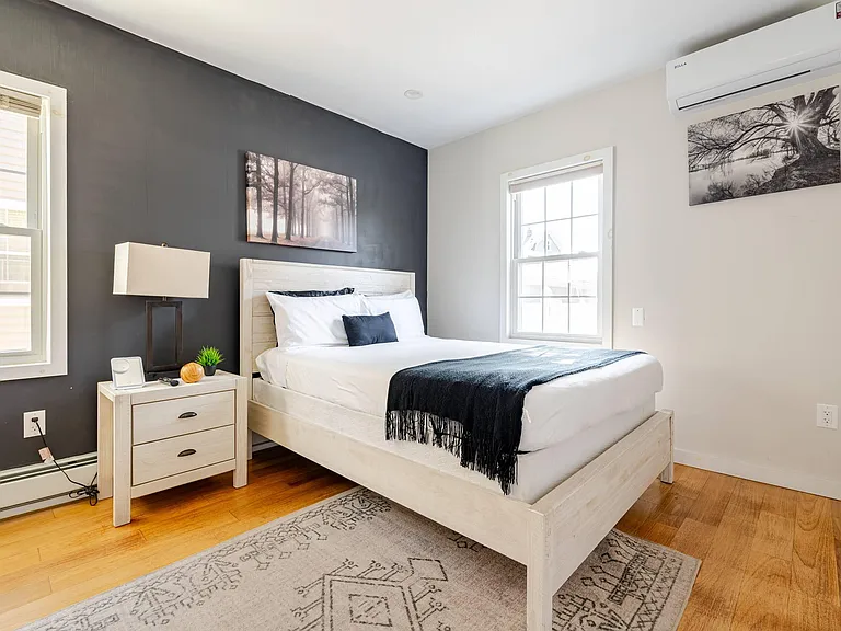 Spacious bedroom with a full bed, nightstand and lamp, hardwood floors, and two windows plus a wall-mounted mini-split.