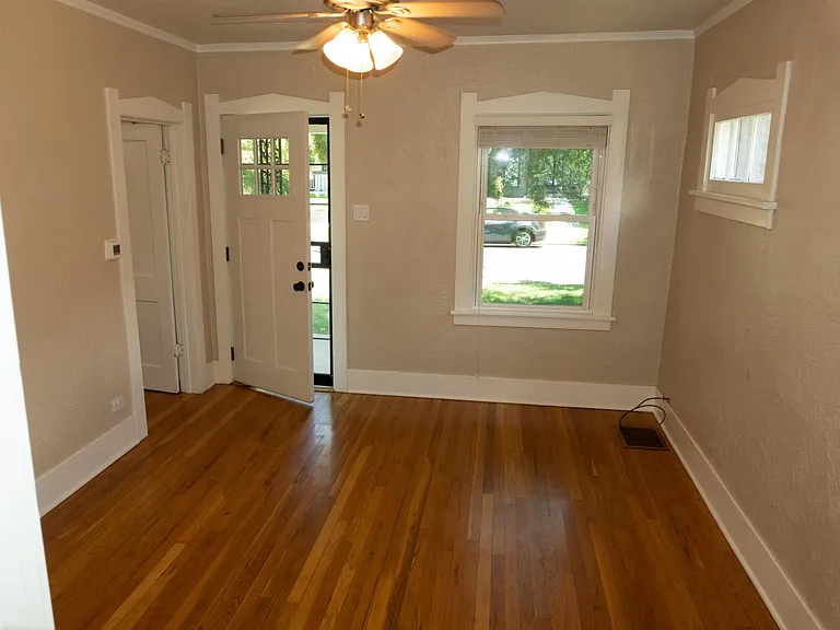 Entering the living room from the kitchen, looking towards the front of the house. The doorway to the front bedroom is left of the front door.