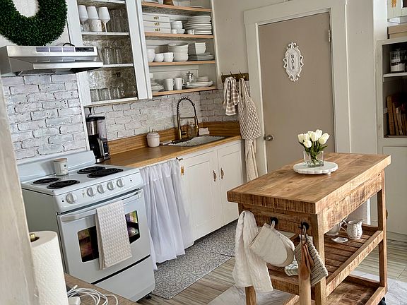 This Scandinavian kitchen combines old-fashioned cottage core design with modern fixtures. The farmhouse style is inviting with open shelving, and linen cabinet coverings bring a cozy feel to this space. (New flooring not shown in this photo)