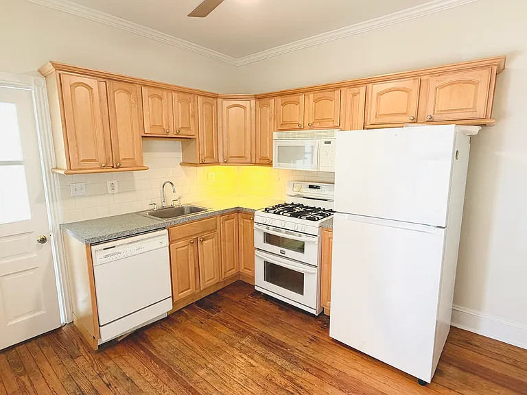 Fully equipped L-shaped kitchen with oak cabinetry, gas stove, dishwasher, microwave, refrigerator, and subway tile backsplash.