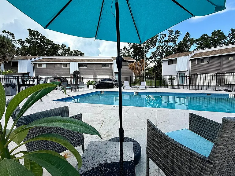 Inviting poolside retreat with chic wicker chairs and a vibrant umbrella, surrounded by modern townhouses and lush greenery.