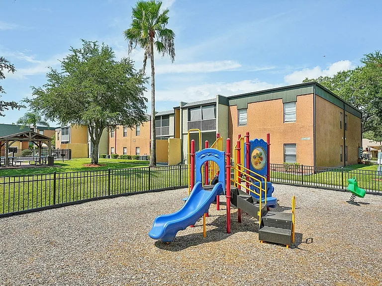 Charming apartment community showcasing a vibrant playground nestled amidst lush greenery and palm trees, perfect for family-friendly living.