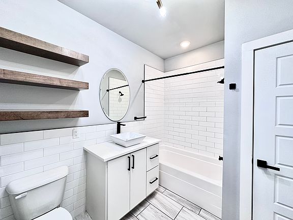 A bathroom that blends modern and rustic charm with statement lighting, tiled backsplash and natural wood shelving for a clean, organic feel. Call or text for a tour today!