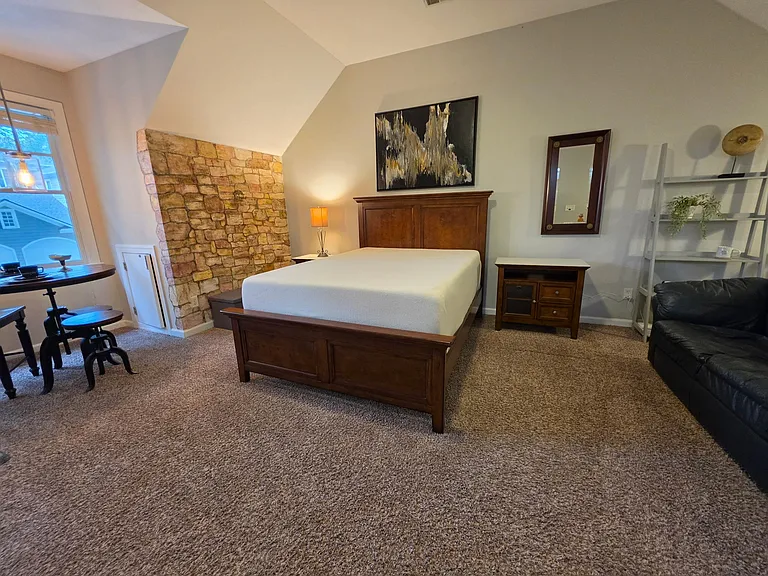 Spacious furnished bedroom with a full bed, nightstand lighting, and a seating nook by the window plus accent stone wall.