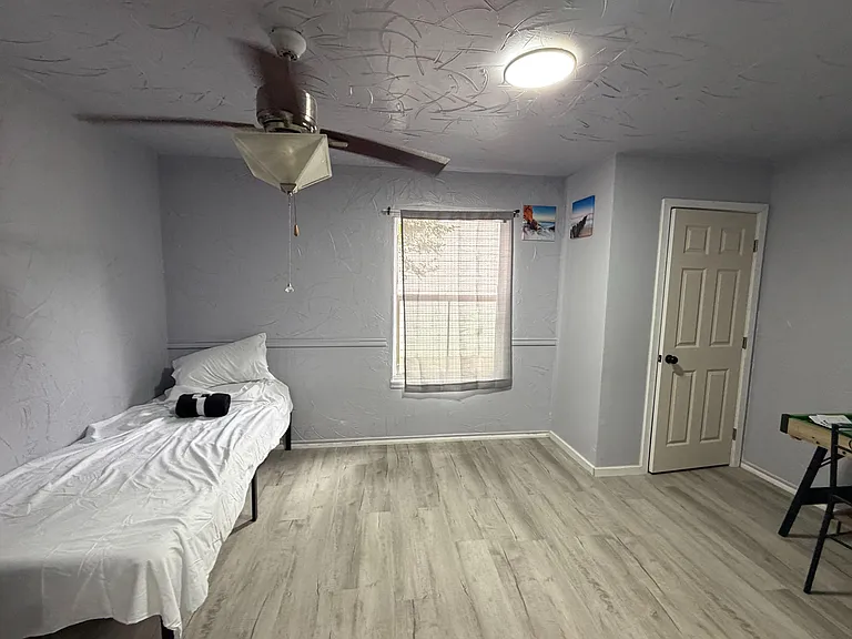 Private bedroom with wood-look flooring, ceiling fan, and a window with blinds; space for a bed and small desk setup.