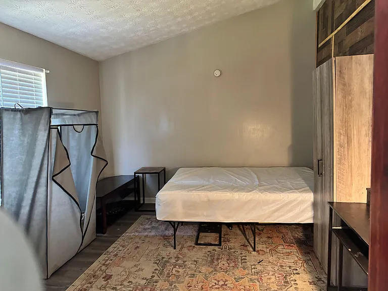 Furnished bedroom with bed, nightstand, and wardrobe, plus a window with blinds and area rug for added comfort.