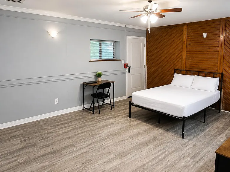 Spacious bedroom with a comfortable bed, dedicated desk workspace, ceiling fan, and durable wood-look flooring.