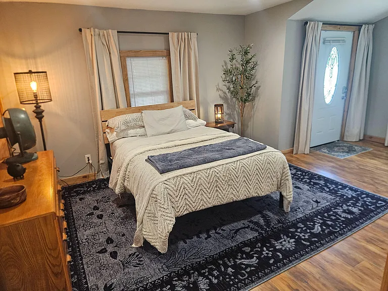 Furnished bedroom with a full bed, bedside tables, dresser, and window treatments, plus durable flooring and a large rug.