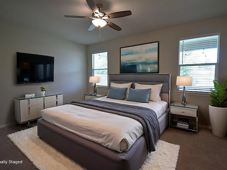 Virtually staged primary bedroom features a ceiling fan and two large windows that provide plenty of natural light