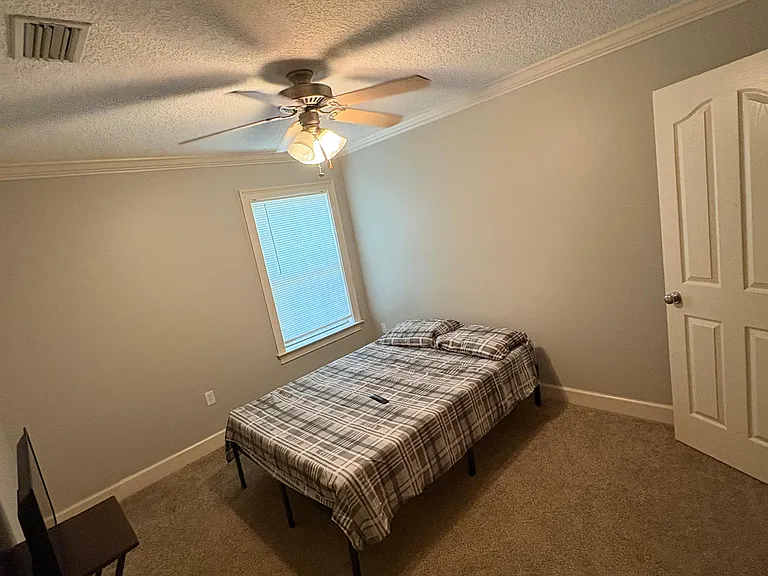 Private bedroom with a ceiling fan, carpeted floors, and a window with blinds, furnished with a bed and small desk area.