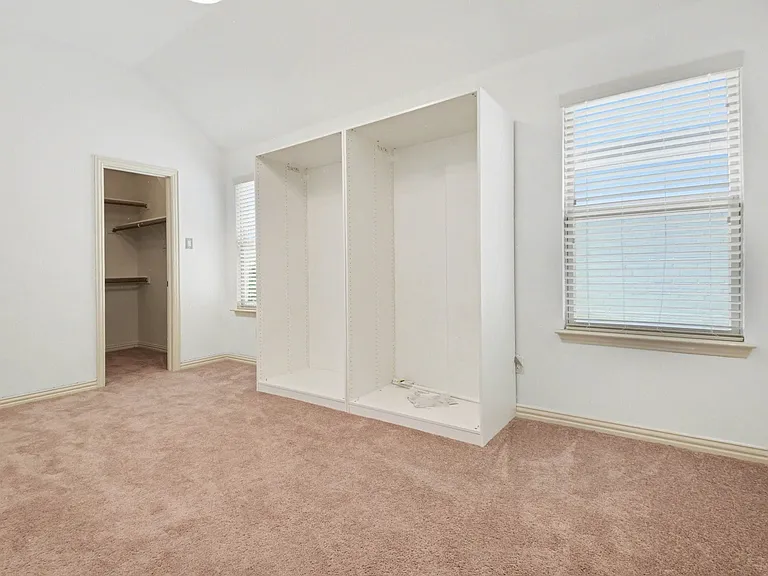 Spacious room featuring vaulted ceilings, plush carpet flooring, two windows with blinds, built-in open shelving, and a walk-in closet with shelving and hanging rods