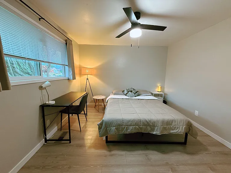 Furnished bedroom with a queen bed, work desk, ceiling fan, and a large window with blinds for natural light.