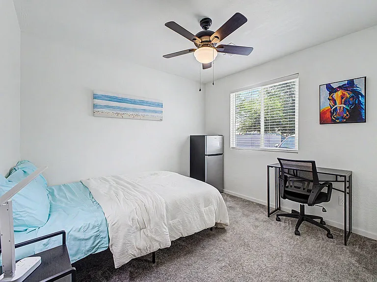 Furnished bedroom with a full bed, dedicated desk setup, ceiling fan, and a large window with blinds for natural light.