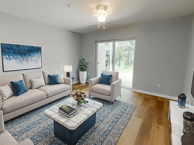 Elegant living room with plush beige sofas, blue accents, a modern marble coffee table, and natural light from large glass doors opening to greenery.