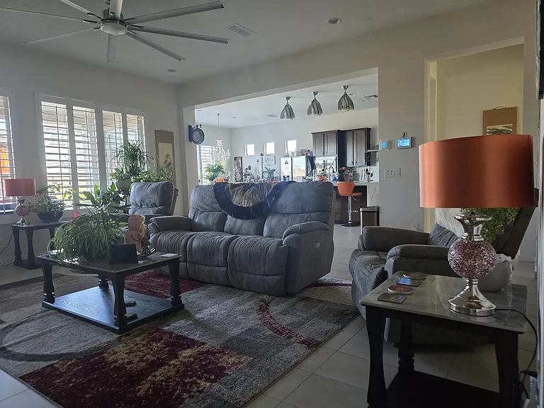 This spacious living room features plush gray recliners, soft patterned carpet, and a glass-top coffee table. Natural light pours through plantation shutters, highlighting potted plants, curated art, and a warm, open layout that flows into the kitchen.