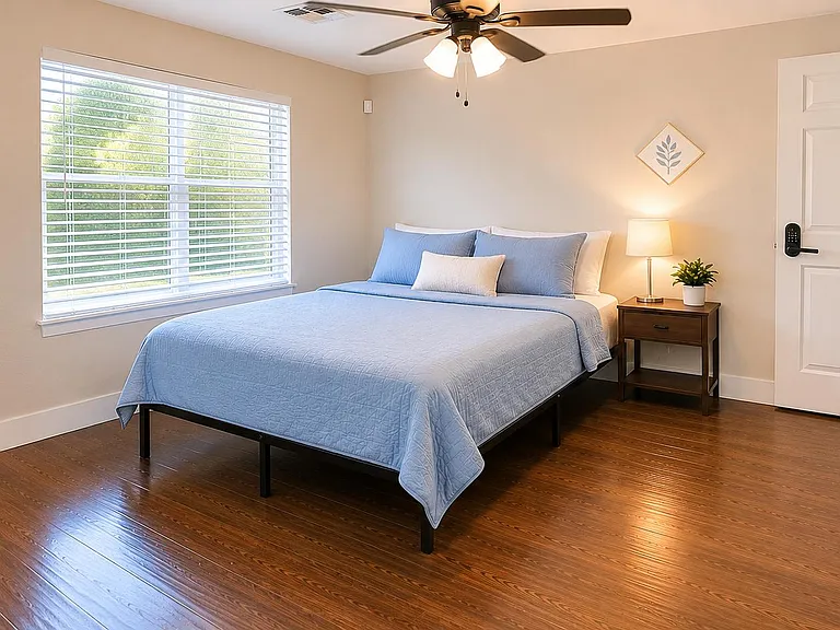 Spacious bedroom with hardwood floors, large window with blinds, ceiling fan, and a nightstand with lamp for easy comfort.