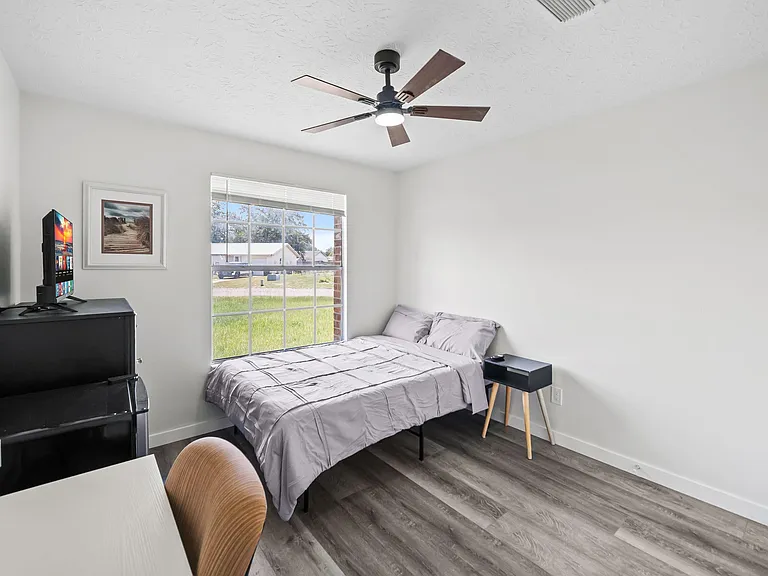 Furnished bedroom with a large window, ceiling fan, and desk area, offering comfortable space for rest and work.