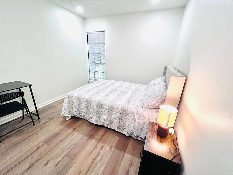 Furnished bedroom with a full bed, nightstand lighting, and a compact desk setup, plus a window with blinds.