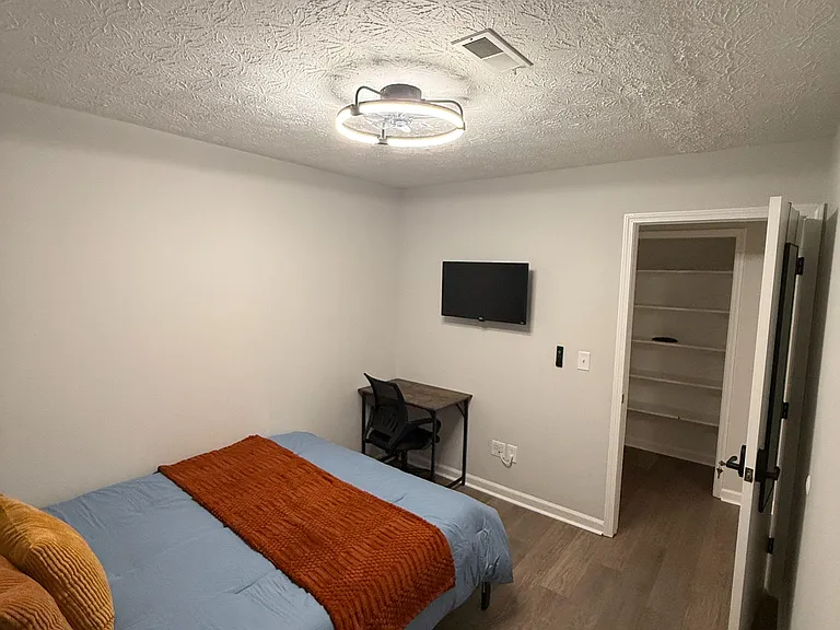 Furnished bedroom with a desk/work area, wall-mounted TV, and an open closet with built-in shelving for storage.