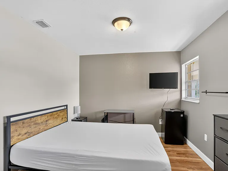 Furnished bedroom with a bed, dresser, wall-mounted TV, and a window for natural light plus a convenient mini fridge.