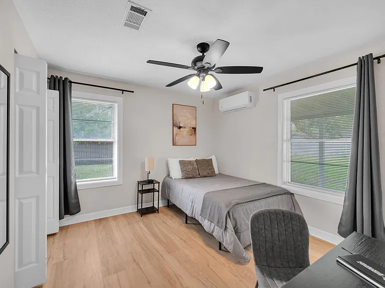 Furnished bedroom with a ceiling fan, wood-look flooring, and two windows with blinds and curtains for privacy.