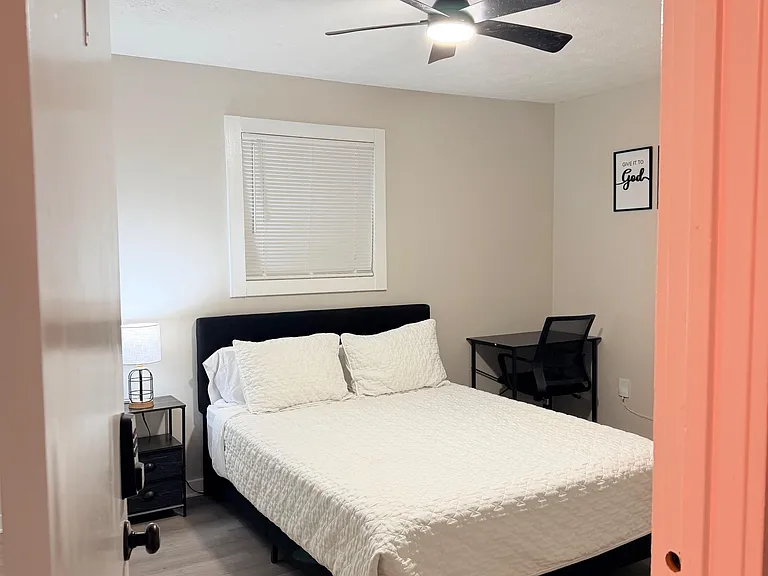 Furnished bedroom with a full bed, nightstand, and dedicated desk space, plus a ceiling fan and window with blinds.