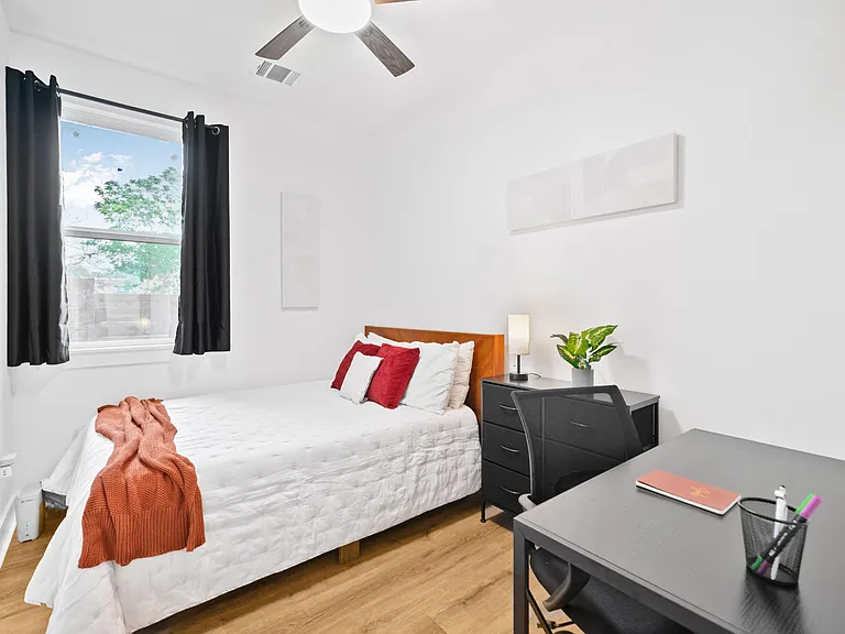 Furnished bedroom with a full bed, nightstand, dresser, and dedicated desk area, plus a large window with curtains.