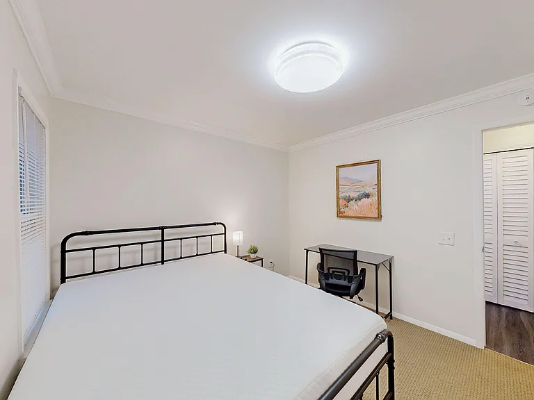 Furnished bedroom with a full-size bed, desk workspace, and closet storage, plus a window with blinds for privacy.