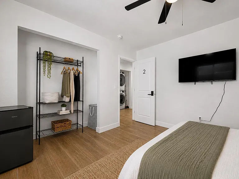 Furnished bedroom with wood-look flooring, ceiling fan, wall-mounted TV, mini fridge, and open closet storage.