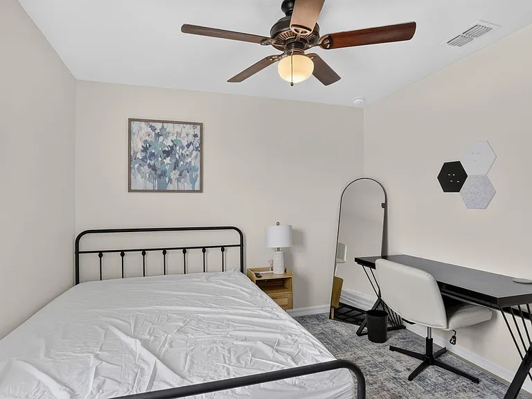 Furnished bedroom with a metal-frame bed, ceiling fan, nightstand, and a dedicated desk/vanity area with mirror.